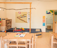 Ein heller, einladender Spielraum für Kinder mit Holzspielzeug und Tischen. In einer Ecke stehen Regale mit Aufbewahrungsboxen. Es gibt eine kleine Kletterfläche mit Teppich, und an der Wand hängen farbige Bilder und ein Informationsblatt.