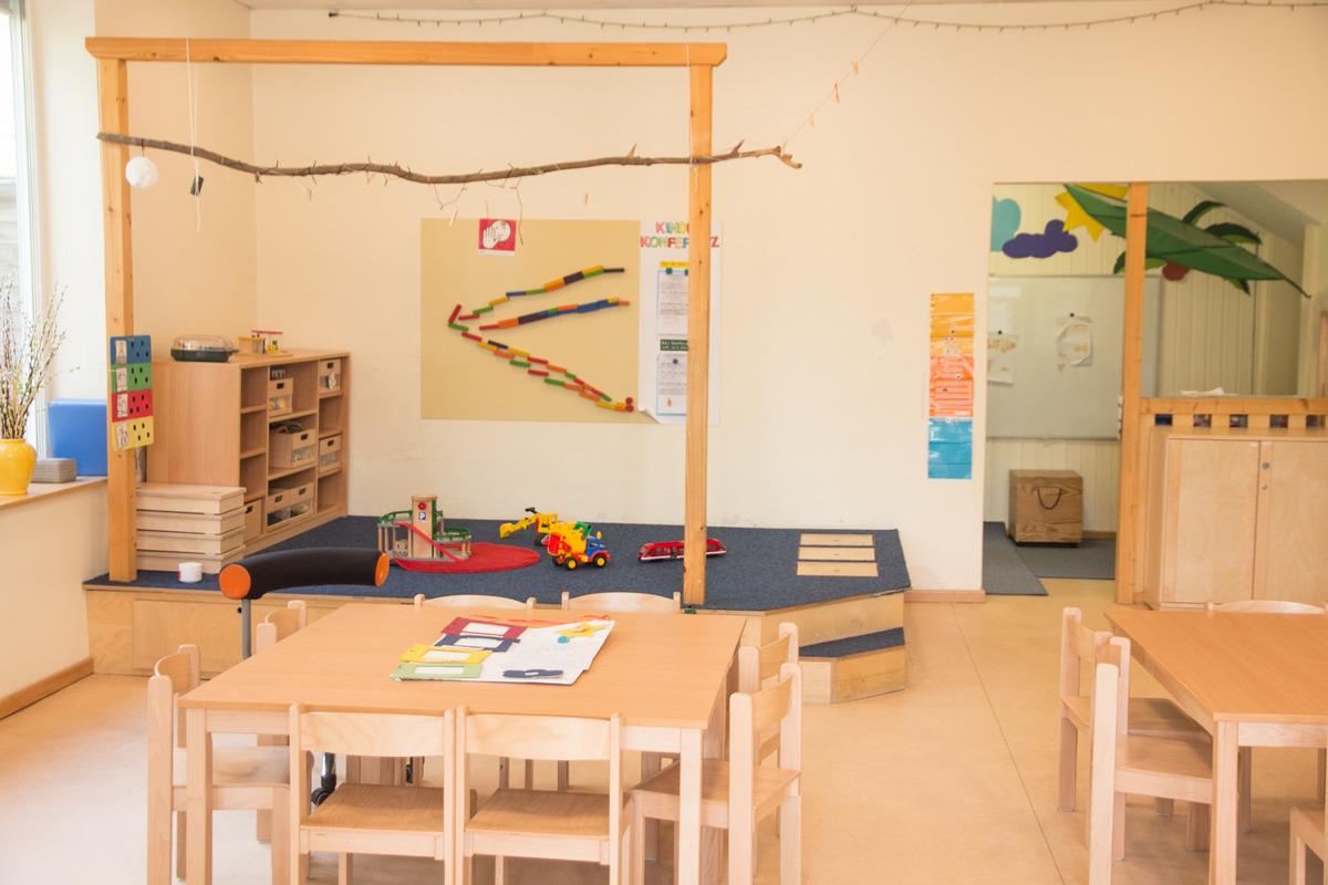 Ein heller, einladender Spielraum für Kinder mit Holzspielzeug und Tischen. In einer Ecke stehen Regale mit Aufbewahrungsboxen. Es gibt eine kleine Kletterfläche mit Teppich, und an der Wand hängen farbige Bilder und ein Informationsblatt.