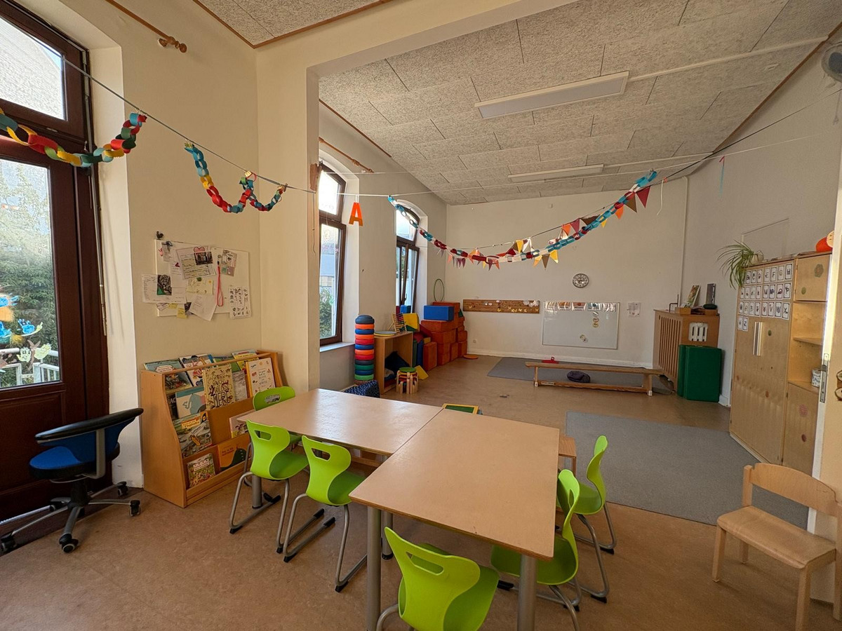 Ein heller Klassenraum mit einem Tisch und grünen Stühlen im Vordergrund. An den Wänden hängen bunte Girlanden. Im Hintergrund befinden sich Stapel von Kissen und ein Lehrtisch. Die Fenster lassen viel Tageslicht herein.