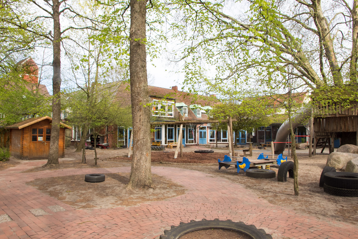 Ein Spielplatz mit mehreren Bäumen und einer im Hintergrund sichtbaren Kita. Es gibt Holzspielgeräte, Schaukeln, eine Rutsche und bunte Holztische. Die Umgebung ist mit Kies und Sand bedeckt, und der Platz ist ansprechend für Kinder gestaltet.