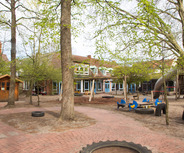 Ein Spielplatz mit mehreren Bäumen und einer im Hintergrund sichtbaren Kita. Es gibt Holzspielgeräte, Schaukeln, eine Rutsche und bunte Holztische. Die Umgebung ist mit Kies und Sand bedeckt, und der Platz ist ansprechend für Kinder gestaltet.