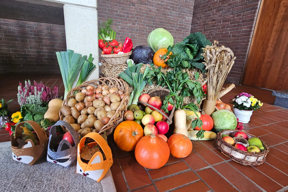 Verschiedene frische Gemüse und Obst sind auf dem Boden angeordnet. Dazu gehören Kürbisse, Äpfel, Kartoffeln, Kohl und Blumenarrangements. Einige Korbgefäße sind sichtbar, ebenso wie dekorative Elemente in Form von Tieren.