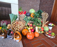Verschiedene frische Gemüse und Obst sind auf dem Boden angeordnet. Dazu gehören Kürbisse, Äpfel, Kartoffeln, Kohl und Blumenarrangements. Einige Korbgefäße sind sichtbar, ebenso wie dekorative Elemente in Form von Tieren.