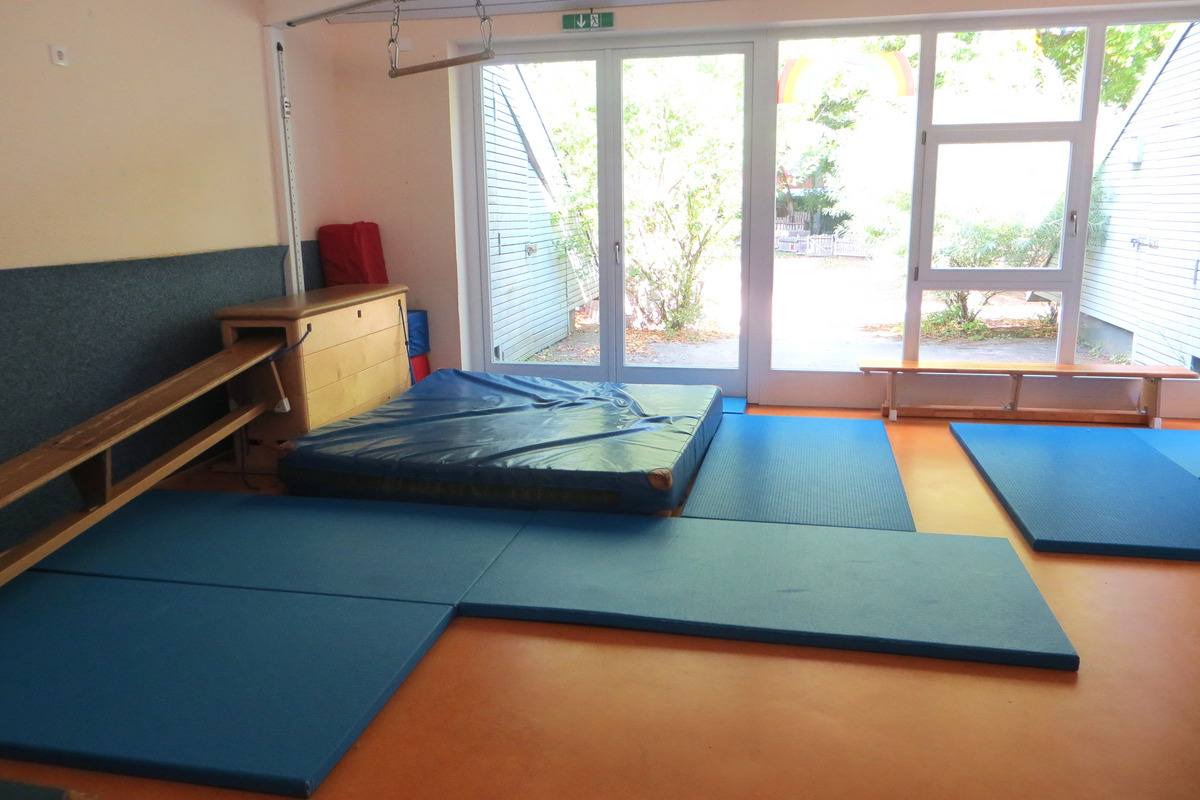 Der Raum zeigt eine Sportfläche mit blauen Matten und einer weichen Landefläche. Ein Fenster öffnet sich nach draußen, wo Pflanzen sichtbar sind. An der Wand stehen eine Holzbank und weitere Matten, die zur Nutzung einladen.
