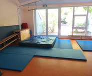 Der Raum zeigt eine Sportfläche mit blauen Matten und einer weichen Landefläche. Ein Fenster öffnet sich nach draußen, wo Pflanzen sichtbar sind. An der Wand stehen eine Holzbank und weitere Matten, die zur Nutzung einladen.