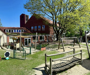 Eine Spielplatzansicht mit mehreren Spielgeräten wie Schaukeln und einer Wippe, umgeben von Rasen und Bäumen. Im Hintergrund gibt es ein Gebäude mit Fenstern und einer strukturierte Dachform. Der Himmel ist blau und es ist ein sonniger Tag.