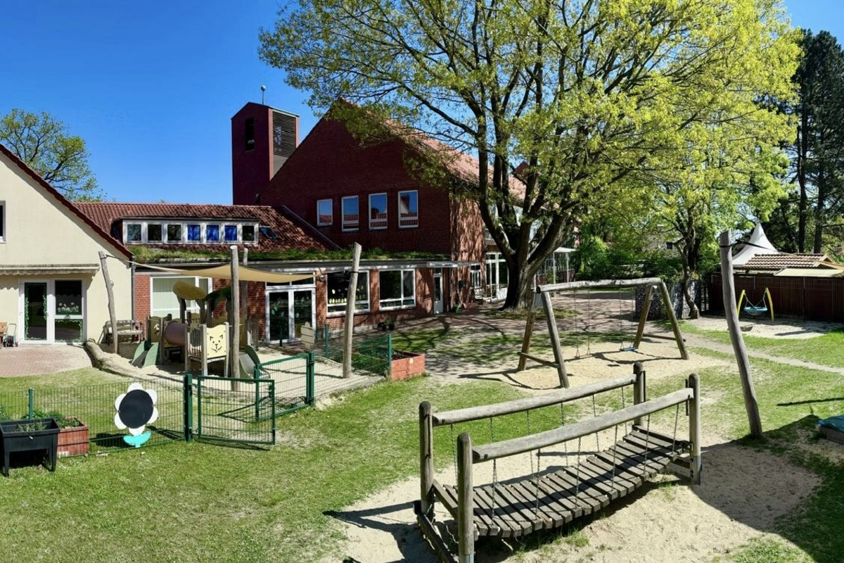 Eine Spielplatzansicht mit mehreren Spielgeräten wie Schaukeln und einer Wippe, umgeben von Rasen und Bäumen. Im Hintergrund gibt es ein Gebäude mit Fenstern und einer strukturierte Dachform. Der Himmel ist blau und es ist ein sonniger Tag.