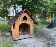 Ein kleines Holzspielhaus steht auf dem Krippenspielplatz. Es hat ein rotes Dach und bunten Wimpel. In der Nähe sind Bäume und ein Spielgerät zu sehen. Der Boden ist sandig.