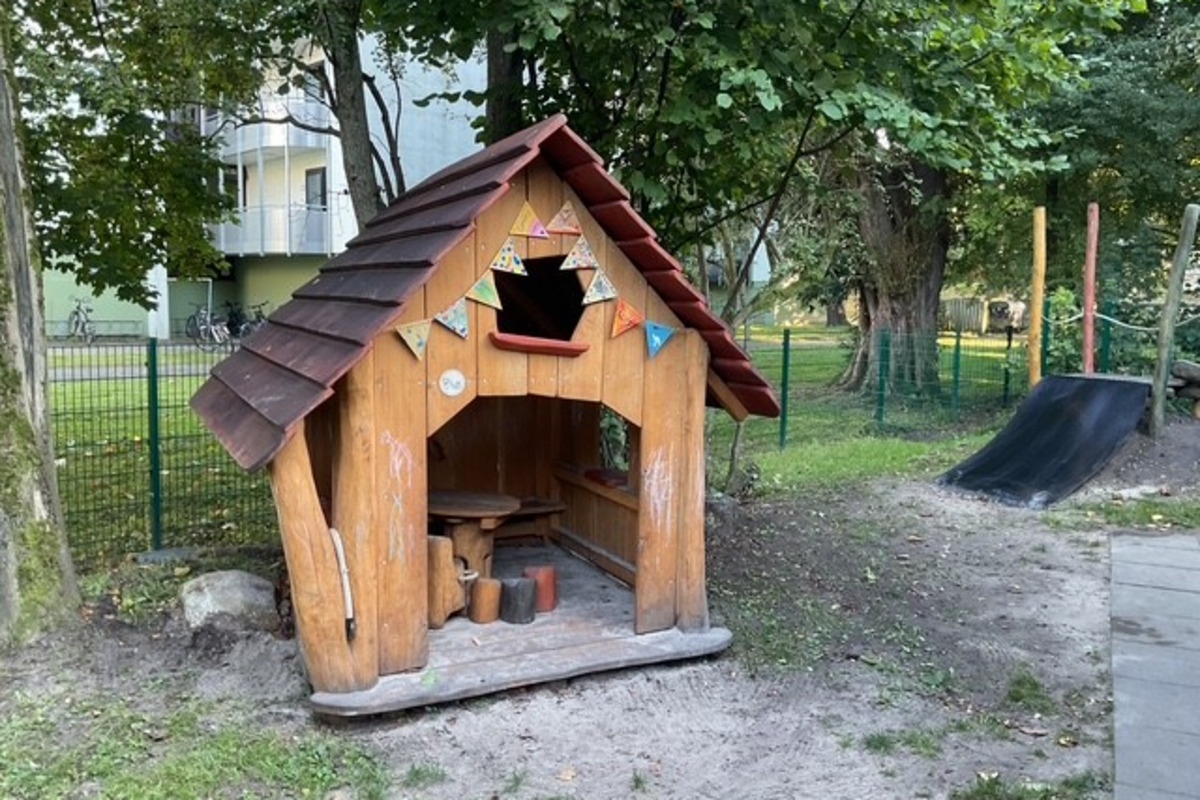 Ein kleines Holzspielhaus steht auf dem Krippenspielplatz. Es hat ein rotes Dach und bunten Wimpel. In der Nähe sind Bäume und ein Spielgerät zu sehen. Der Boden ist sandig.