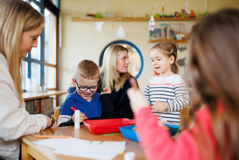 In einem hellen Raum stehen zwei Kinder an einem Tisch und spielen mit Bastelmaterial. Ein Junge mit Brille zeichnet eifrig, während ein Mädchen aufgeregt spricht. Im Hintergrund sind Erwachsene zu sehen, die die Kinder beobachten und unterstützen.