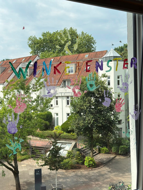Blick aus einem Fenster auf einen Garten. Auf dem Fenster sind bunte Handabdrücke und die Worte "WINKFENSTER" zu sehen. Im Hintergrund stehen Bäume und Gebäude mit roten Dächern.