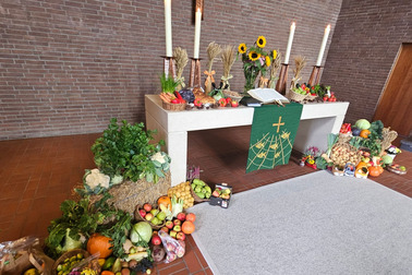 Ein festlich gedeckter Altar mit einer Vielzahl von Obst und Gemüse. Auf dem Tisch stehen eine Bibel, drei Kerzen und Sonnenblumen. Um den Altar liegen weitere Früchte und Gemüse, die eine Erntedankatmosphäre schaffen. Im Hintergrund eine Steinwand.