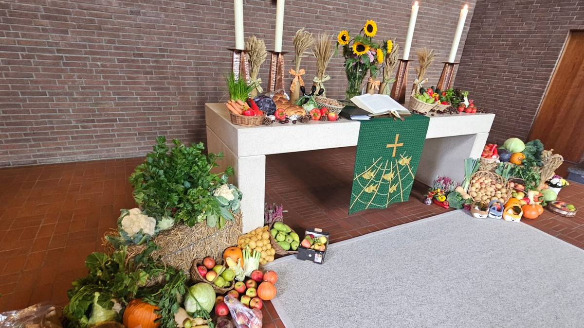 Ein festlich gedeckter Altar mit einer Vielzahl von Obst und Gemüse. Auf dem Tisch stehen eine Bibel, drei Kerzen und Sonnenblumen. Um den Altar liegen weitere Früchte und Gemüse, die eine Erntedankatmosphäre schaffen. Im Hintergrund eine Steinwand.