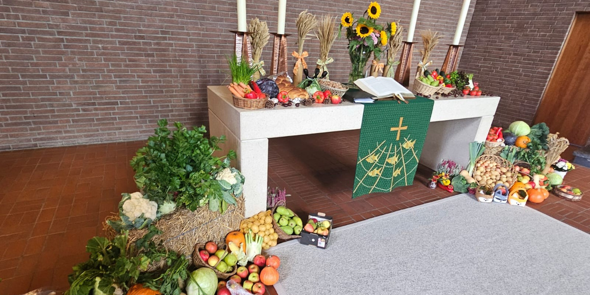 Ein festlich gedeckter Altar mit einer Vielzahl von Obst und Gemüse. Auf dem Tisch stehen eine Bibel, drei Kerzen und Sonnenblumen. Um den Altar liegen weitere Früchte und Gemüse, die eine Erntedankatmosphäre schaffen. Im Hintergrund eine Steinwand.