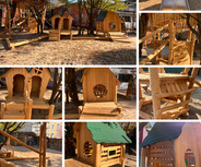Das Bild zeigt einen Spielplatz mit Holzspielgeräten, darunter mehrere kleine Spielhäuser, Rutschen und Kletterelemente. 