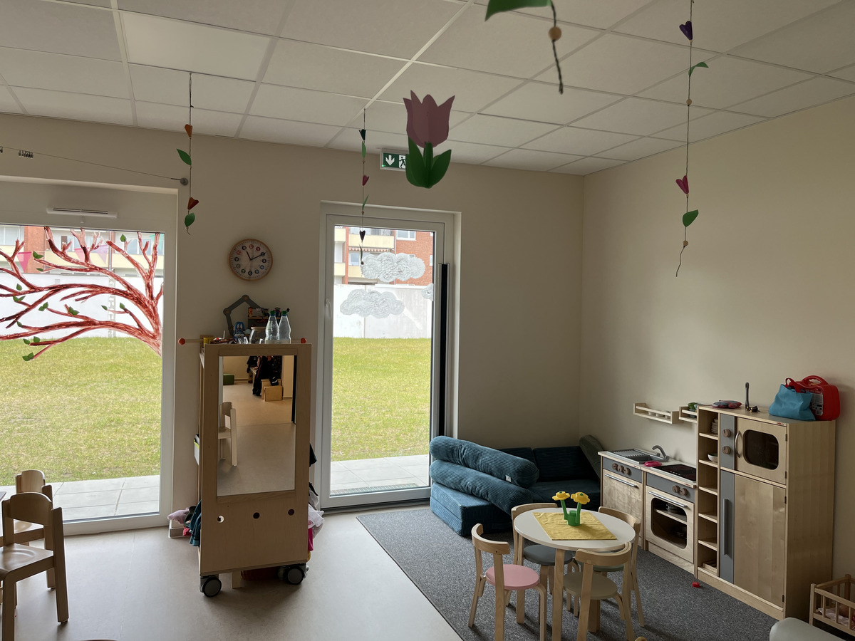 Der Raum ist ein helles Spielzimmer mit einem runden Tisch, umgeben von kleinen Stühlen. An der Wand gibt es eine farbenfrohe Baummalerei. Ein Sofa steht in der Ecke, und ein Spielbereich mit einer kleinen Küche ist sichtbar. Fenster lassen Tageslicht herein.