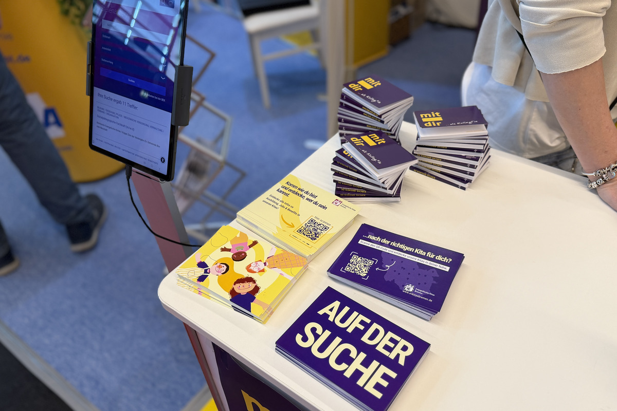 Ein Messestand mit einem Tisch, auf dem stapelbare Karten mit dem Text "AUF DER SUCHE" liegen. Daneben befindet sich ein QR-Code und ein Tablet, das möglicherweise Informationen anzeigt. 
