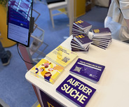 Ein Messestand mit einem Tisch, auf dem stapelbare Karten mit dem Text "AUF DER SUCHE" liegen. Daneben befindet sich ein QR-Code und ein Tablet, das möglicherweise Informationen anzeigt. 