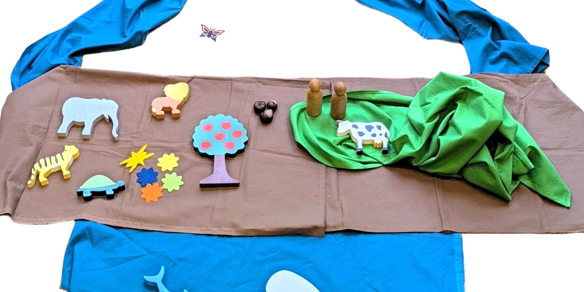 Eine Spielszenen mit bunten Holzfiguren, darunter Tiere wie ein Elefant und ein Zebra, sowie eine Pflanze und eine Kuh. Das Arrangement umfasst ein blaues Tuch im Hintergrund und grüne Stoffelemente, die eine Landschaft darstellen.