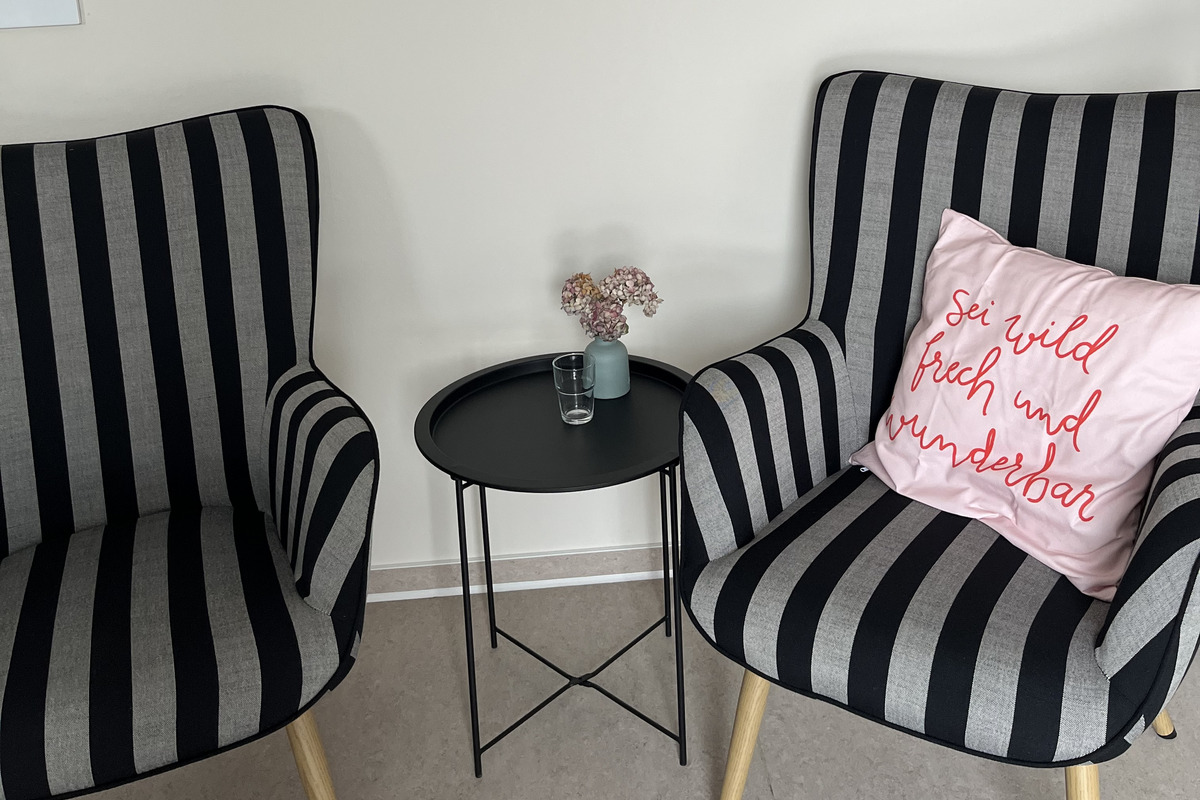 Zwei schwarz-weiß gestreifte Sessel stehen sich gegenüber. Auf dem kleinen schwarzen Tisch zwischen ihnen steht eine Vase mit kleinen Blumen und ein Glas. An der Wand hängt ein abstraktes Kunstwerk in sanften Rosa- und Rottönen. Ein Kissen mit der Aufschrift „sei wild, frisch und wunderbar“ liegt auf einem der Sessel.
