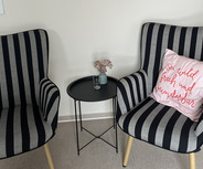 Zwei schwarz-weiß gestreifte Sessel stehen sich gegenüber. Auf dem kleinen schwarzen Tisch zwischen ihnen steht eine Vase mit kleinen Blumen und ein Glas. An der Wand hängt ein abstraktes Kunstwerk in sanften Rosa- und Rottönen. Ein Kissen mit der Aufschrift „sei wild, frisch und wunderbar“ liegt auf einem der Sessel.