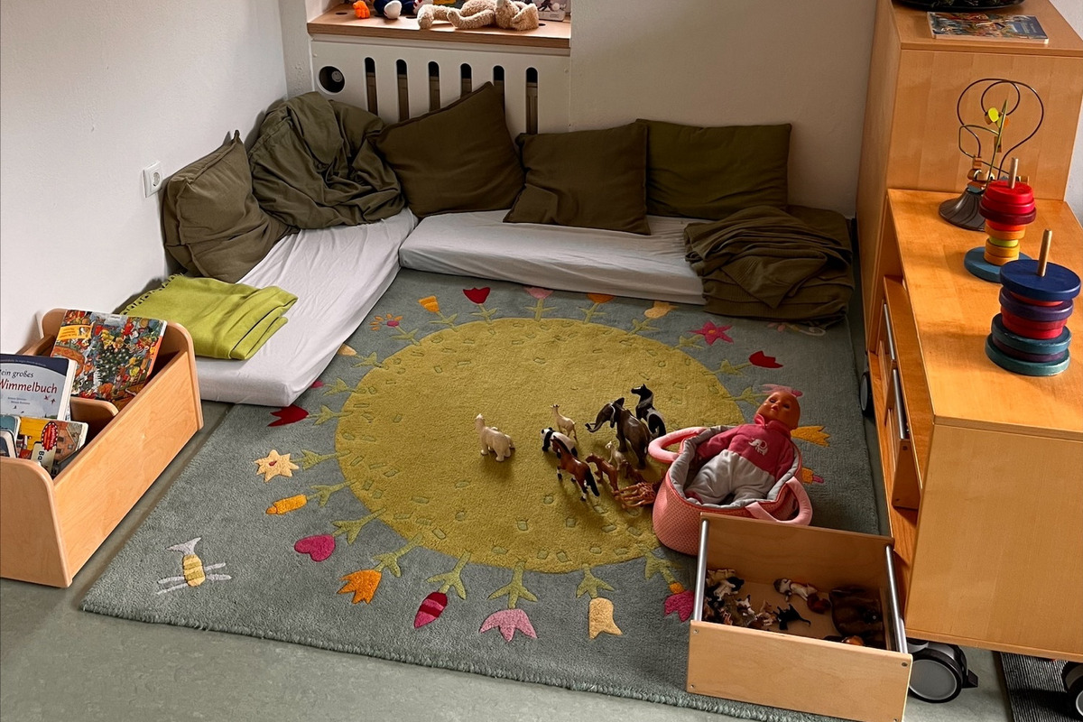 Der Raum zeigt einen Spielbereich mit einem bunten Teppich, auf dem Spielzeug wie Figuren und eine Puppe liegen. 