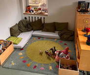 Der Raum zeigt einen Spielbereich mit einem bunten Teppich, auf dem Spielzeug wie Figuren und eine Puppe liegen. 