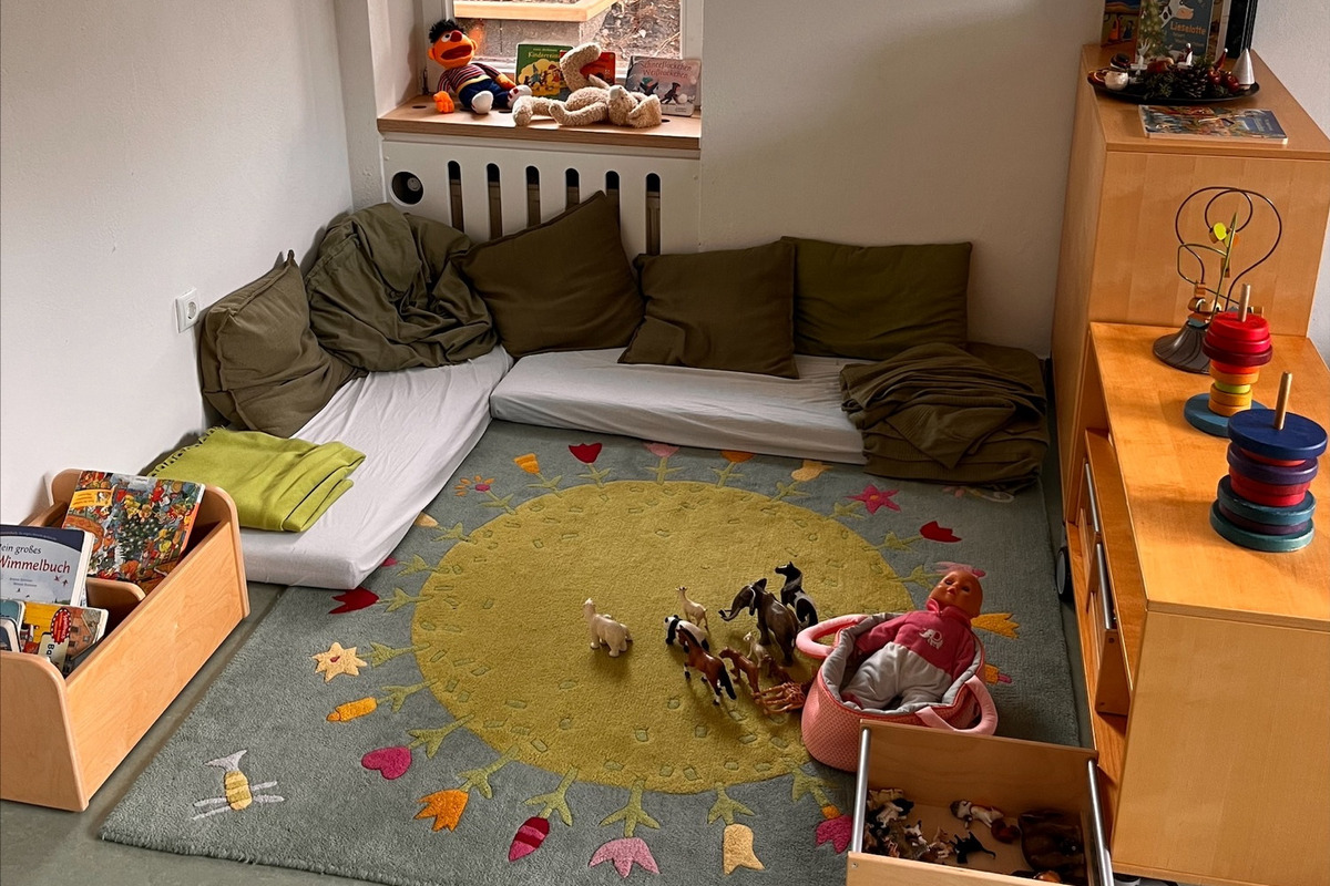 Der Raum zeigt einen Spielbereich mit einem bunten Teppich, auf dem Spielzeug wie Figuren und eine Puppe liegen. 