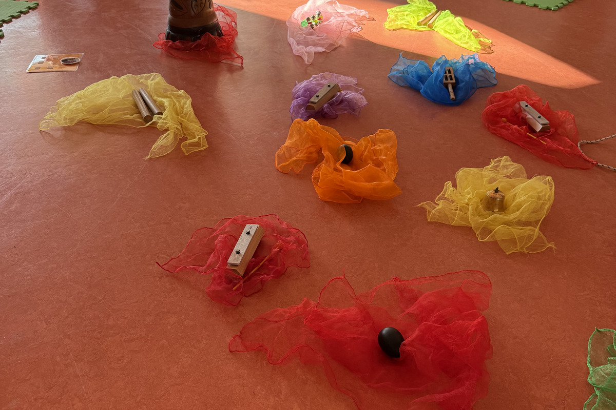 Im Raum liegen mehrere farbige Tücher in Rot, Gelb, Blau, Orange und Lila, die mit verschiedenen Musikinstrumenten wie Klanghölzern und einem Arbeitsplatz aus einem Trommel stehen.