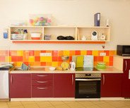 Eine moderne Küche für Kinder mit einer Kombination aus roten und orangefarbenen Kacheln. Die Arbeitsfläche enthält einen Wasserhahn, eine Spüle, eine Küchenwaage und einen Mixer. Oben sind verschiedene Aufbewahrungsbehälter und Küchengeräte angeordnet, darunter eine Mikrowelle und ein Ofen.