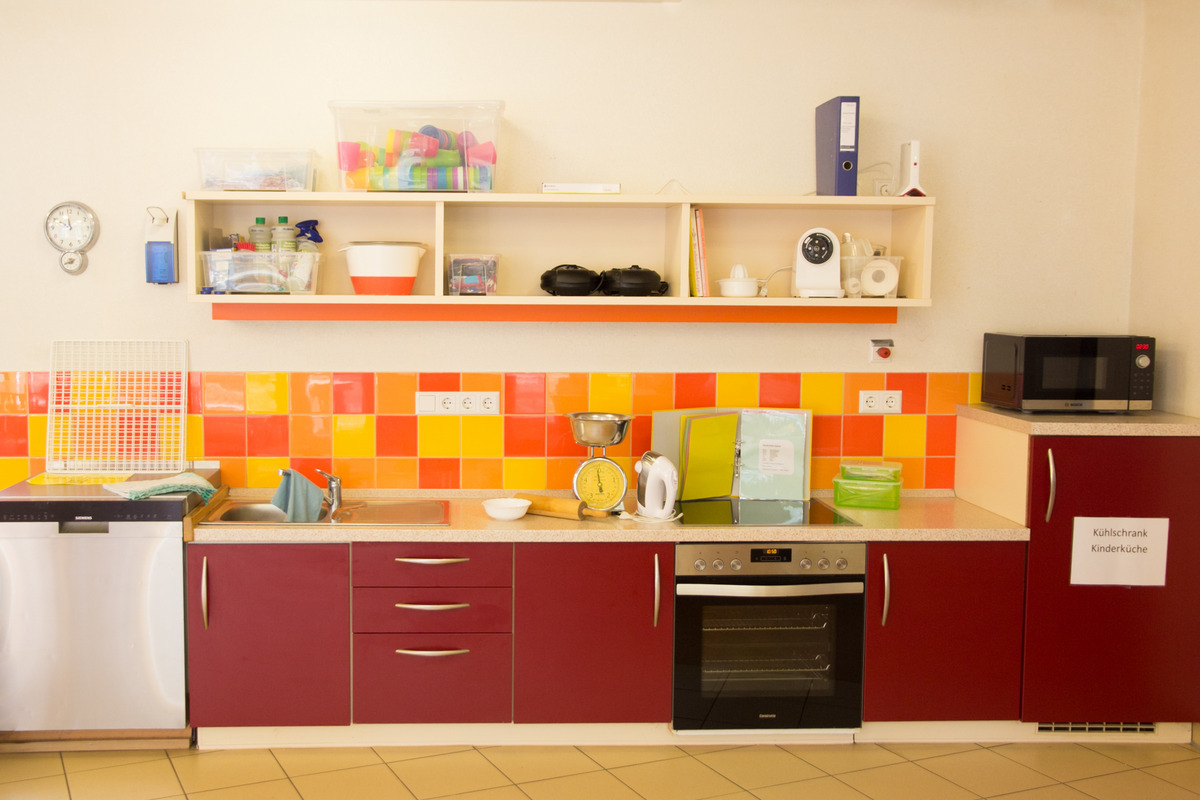 Eine moderne Küche für Kinder mit einer Kombination aus roten und orangefarbenen Kacheln. Die Arbeitsfläche enthält einen Wasserhahn, eine Spüle, eine Küchenwaage und einen Mixer. Oben sind verschiedene Aufbewahrungsbehälter und Küchengeräte angeordnet, darunter eine Mikrowelle und ein Ofen.