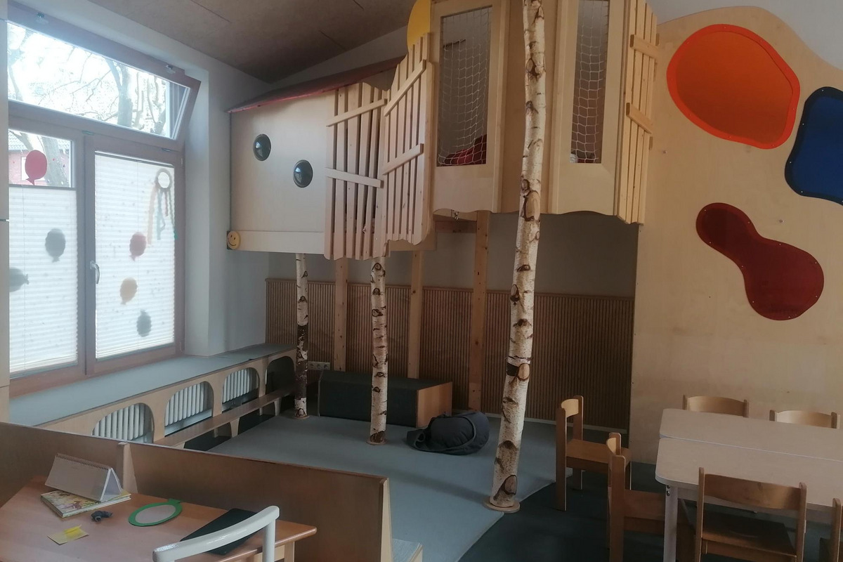 Der Raum ist ein Spielzimmer mit einem erhöhten Holzspielhaus, in das drei Baumstämme integriert sind. 