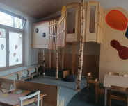 Der Raum ist ein Spielzimmer mit einem erhöhten Holzspielhaus, in das drei Baumstämme integriert sind. 