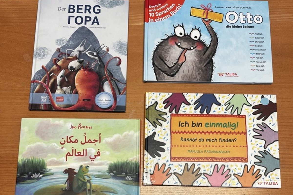 Auf einem Holztisch liegen sechs Kinderbücher. Die Titel sind in verschiedenen Sprachen und beinhalten Themen wie Freundschaft, Farben und Geschichten. Farbige Illustrationen und unterschiedliche Schriftarten sind sichtbar.