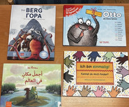 Auf einem Holztisch liegen sechs Kinderbücher. Die Titel sind in verschiedenen Sprachen und beinhalten Themen wie Freundschaft, Farben und Geschichten. Farbige Illustrationen und unterschiedliche Schriftarten sind sichtbar.