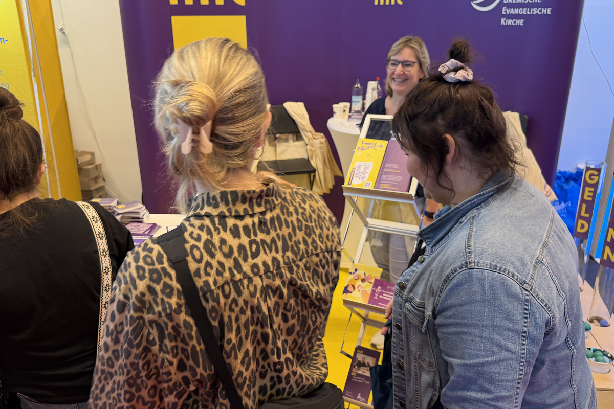 Eine Gruppe von Menschen steht an einem Messestand. Die Personen sind rückseitig zu sehen. Eine Frau an dem Stand lächelt und präsentiert Informationsmaterial. Im Hintergrund sind gelbe Banner mit dem Schriftzug „GELD“, „SINN“ und „SPASS“ zu erkennen.