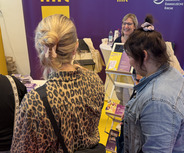 Eine Gruppe von Menschen steht an einem Messestand. Die Personen sind rückseitig zu sehen. Eine Frau an dem Stand lächelt und präsentiert Informationsmaterial. Im Hintergrund sind gelbe Banner mit dem Schriftzug „GELD“, „SINN“ und „SPASS“ zu erkennen.
