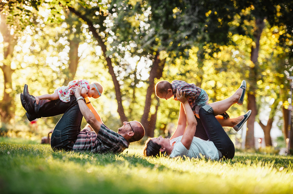 Eine Familie mit zwei kleinen Kindern spielt im Park. Der Vater liegt auf dem Rücken und hebt seine Tochter in die Luft, während die Mutter ihren Sohn ebenfalls anhebt. Alle lachen und genießen die Zeit im Freien, umgeben von Bäumen und Sonnenschein.