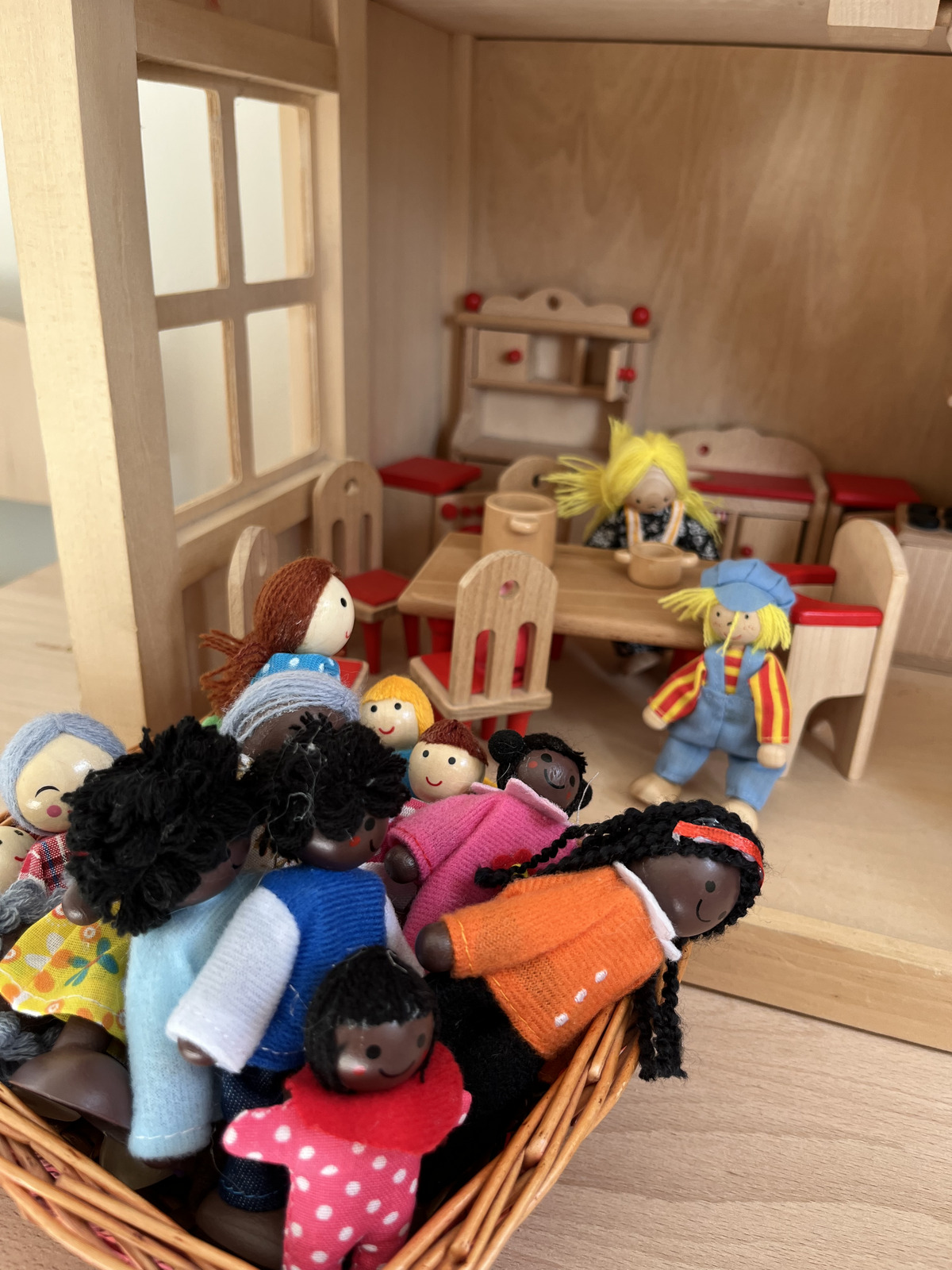 In einem Spielzeughaus sind zahlreiche bunte Puppen versammelt. Im Vordergrund befinden sich viele Puppen in einem Korb. Im Hintergrund sitzen einige Puppen an einem Tisch, während zwei Puppen spielend aufstehen. Das Szenario vermittelt eine fröhliche, interaktive Spielatmosphäre.