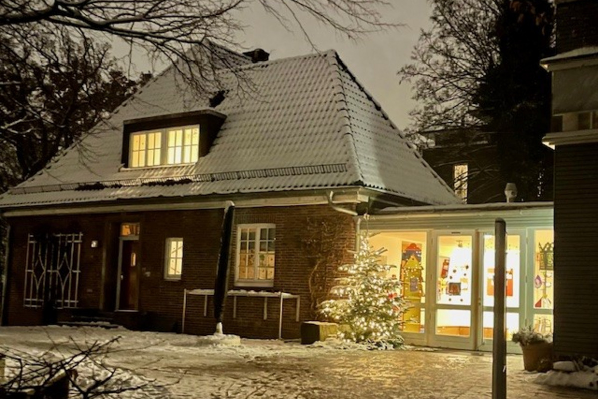 Ein hell erleuchtetes Gebäude mit einem schneebedeckten Dach. Vor dem Eingang steht ein Weihnachtsbaum. Der Boden ist mit Schnee bedeckt und die Bäume im Hintergrund sind teilweise kahl. Die Atmosphäre wirkt gemütlich und winterlich.