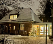 Ein hell erleuchtetes Gebäude mit einem schneebedeckten Dach. Vor dem Eingang steht ein Weihnachtsbaum. Der Boden ist mit Schnee bedeckt und die Bäume im Hintergrund sind teilweise kahl. Die Atmosphäre wirkt gemütlich und winterlich.