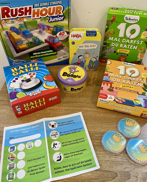 Auf einem Tisch liegen verschiedene Gesellschaftsspiele für Kinder. Dazu gehören "Rush Hour Junior", "Halli Galli", "Dobble" und "Wer bin ich?". Außerdem befindet sich ein Flyer mit Hinweisen zur Spielnutzung und Belohnungen für das Spielen.