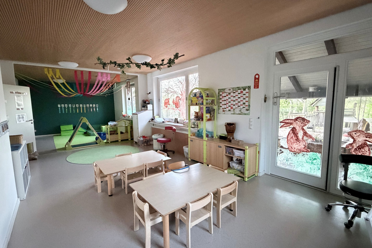 Innenansicht eines Kindergartens mit mehreren Tischen und Stühlen. An der Wand hängen farbige Dekorationen, und ein grüner Bereich mit Spielgeräten ist sichtbar. Fenster bieten einen Blick nach draußen. Der Raum ist freundlich und einladend gestaltet.