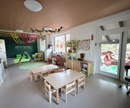 Innenansicht eines Kindergartens mit mehreren Tischen und Stühlen. An der Wand hängen farbige Dekorationen, und ein grüner Bereich mit Spielgeräten ist sichtbar. Fenster bieten einen Blick nach draußen. Der Raum ist freundlich und einladend gestaltet.