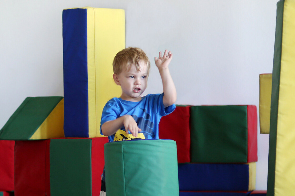 Ein kleiner Junge in einem blauen T-Shirt steht zwischen bunten Schaumstoffblöcken. Er hebt eine Hand und hält ein gelbes Spielzeug in der anderen. Die Blöcke sind in verschiedenen Farben angeordnet, darunter Grün, Rot und Blau.