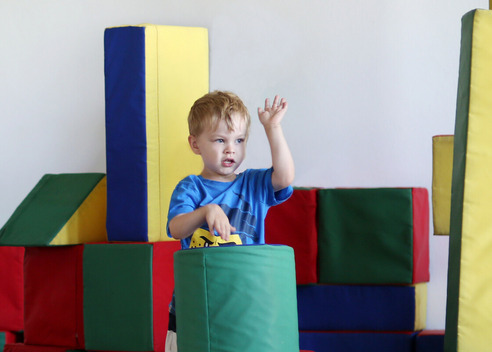 Ein kleiner Junge in einem blauen T-Shirt steht zwischen bunten Schaumstoffblöcken. Er hebt eine Hand und hält ein gelbes Spielzeug in der anderen. Die Blöcke sind in verschiedenen Farben angeordnet, darunter Grün, Rot und Blau.
