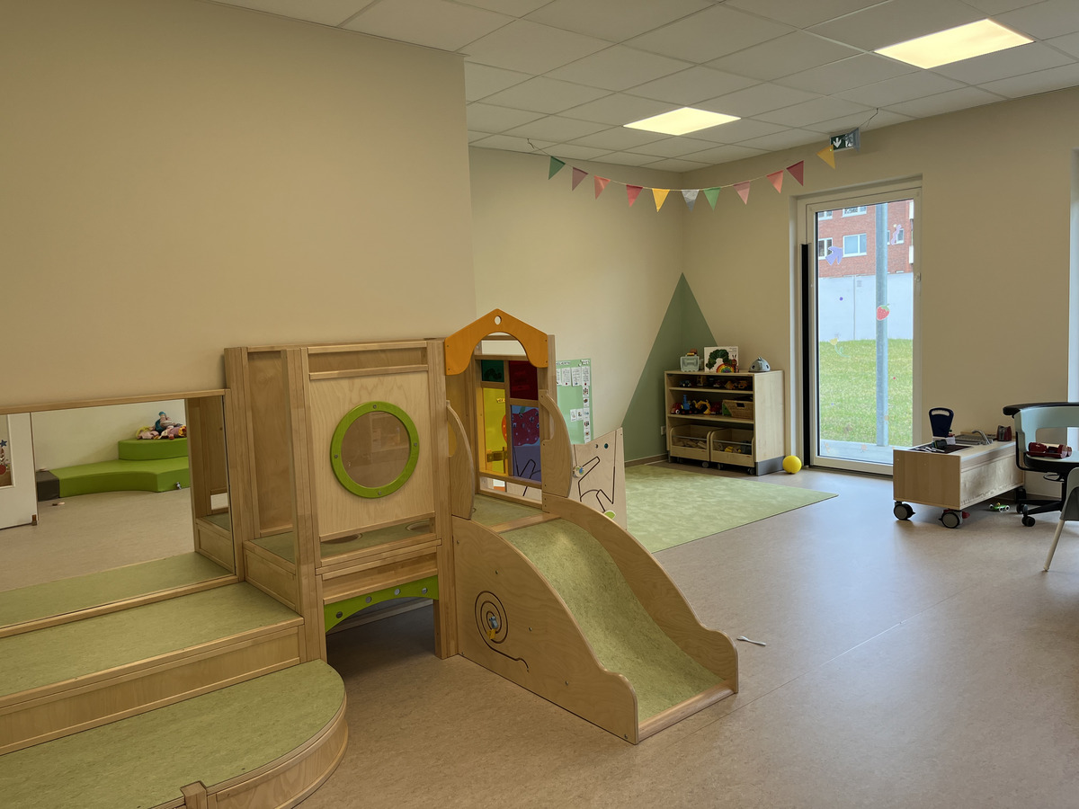 Ein heller Kindergartenraum mit einem Holzspielgerät, das eine Rutsche und einen Spielbereich umfasst. Im Hintergrund stehen ein Regal mit Spielzeug und ein grünes Sofa. Große Fenster bieten Blick auf einen Außenbereich mit Gras. Bunte Wimpel sind an der Decke angebracht.