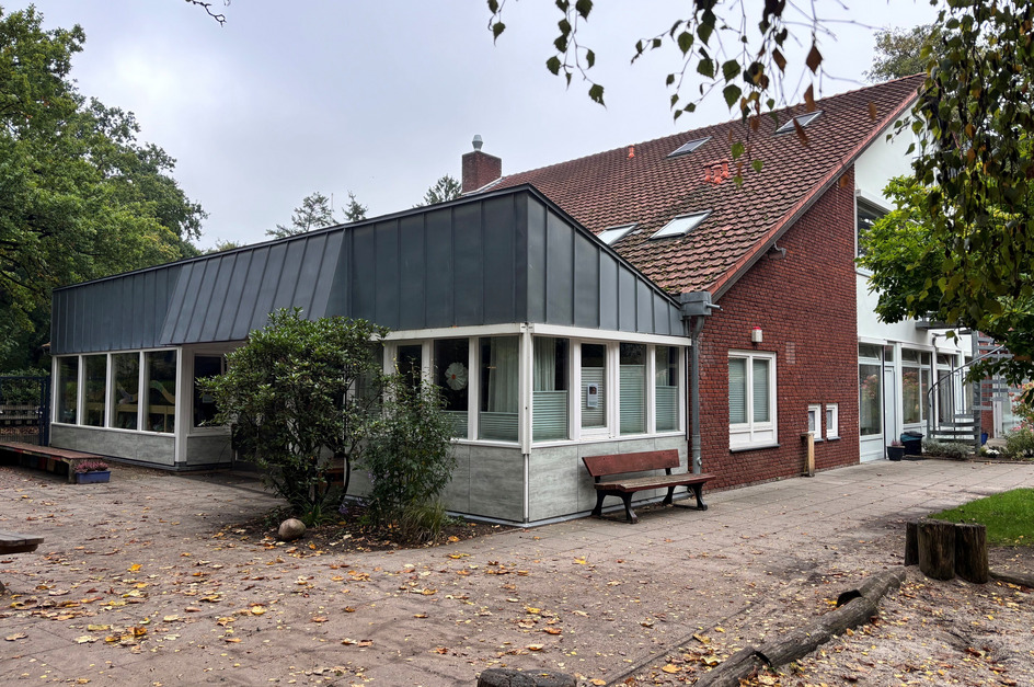 Das Bild zeigt ein modernes Gebäude mit Glaselementen und einem flachen Dach, umgeben von Bäumen und einem befestigten Weg. Vor dem Gebäude stehen eine Bank und einige Pflanzen. 