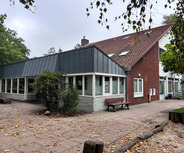 Das Bild zeigt ein modernes Gebäude mit Glaselementen und einem flachen Dach, umgeben von Bäumen und einem befestigten Weg. Vor dem Gebäude stehen eine Bank und einige Pflanzen. 