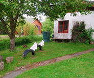 Ein kleines Wohnhaus mit roten Fensterrahmen steht in einem Garten. Vor dem Haus gibt es eine Rasenfläche mit Holzscheiten und zwei weißen, stilisierten Figuren. Im Hintergrund sind grüne Bäume und ein Mülltonne sichtbar. Der Weg zum Haus ist gepflastert.